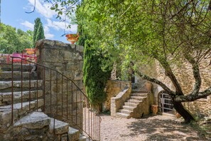 Un escalier en pierre mène à un bâtiment doté d’une rambarde en métal, le tout étant entouré de verdure et surplombé par un ciel dégagé.