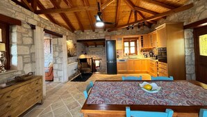 A rustic interior with a stone wall, wooden ceiling beams, a dining table with chairs, a kitchen area, and a cozy seating area. - Villa Ionis Holiday Villa (Volimes)