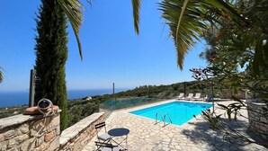 A stone pool with clear blue water, surrounded by stone walls and palm trees, overlooking a scenic landscape. - Villa Ionis Holiday Villa (Volimes)