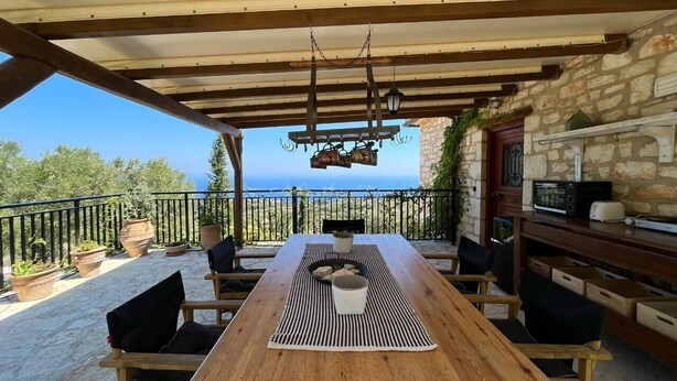 A wooden table with chairs under a pergola, overlooking a stone building and the sea. - Villa Ionis Holiday Villa (Volimes)