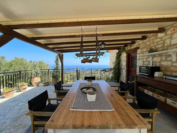Ein Holztisch mit Stühlen unter einer Pergola, mit Blick auf ein Steinhaus und das Meer.