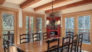 A dining area with a wooden table and chairs, large windows, and a chandelier.