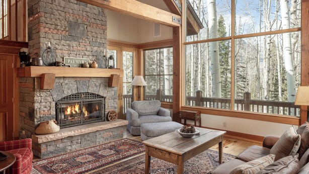 A cozy living room with a stone fireplace, large windows, and a view of trees.