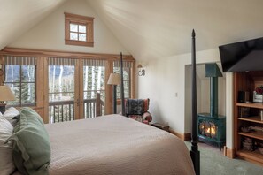 A bedroom with a large bed, a wooden ceiling, a fireplace, a TV, and a view of trees through the windows. - Luxury Telluride Mountain Village Home | Hot Tub, Fireplace, Sleeps 12 (Telluride)