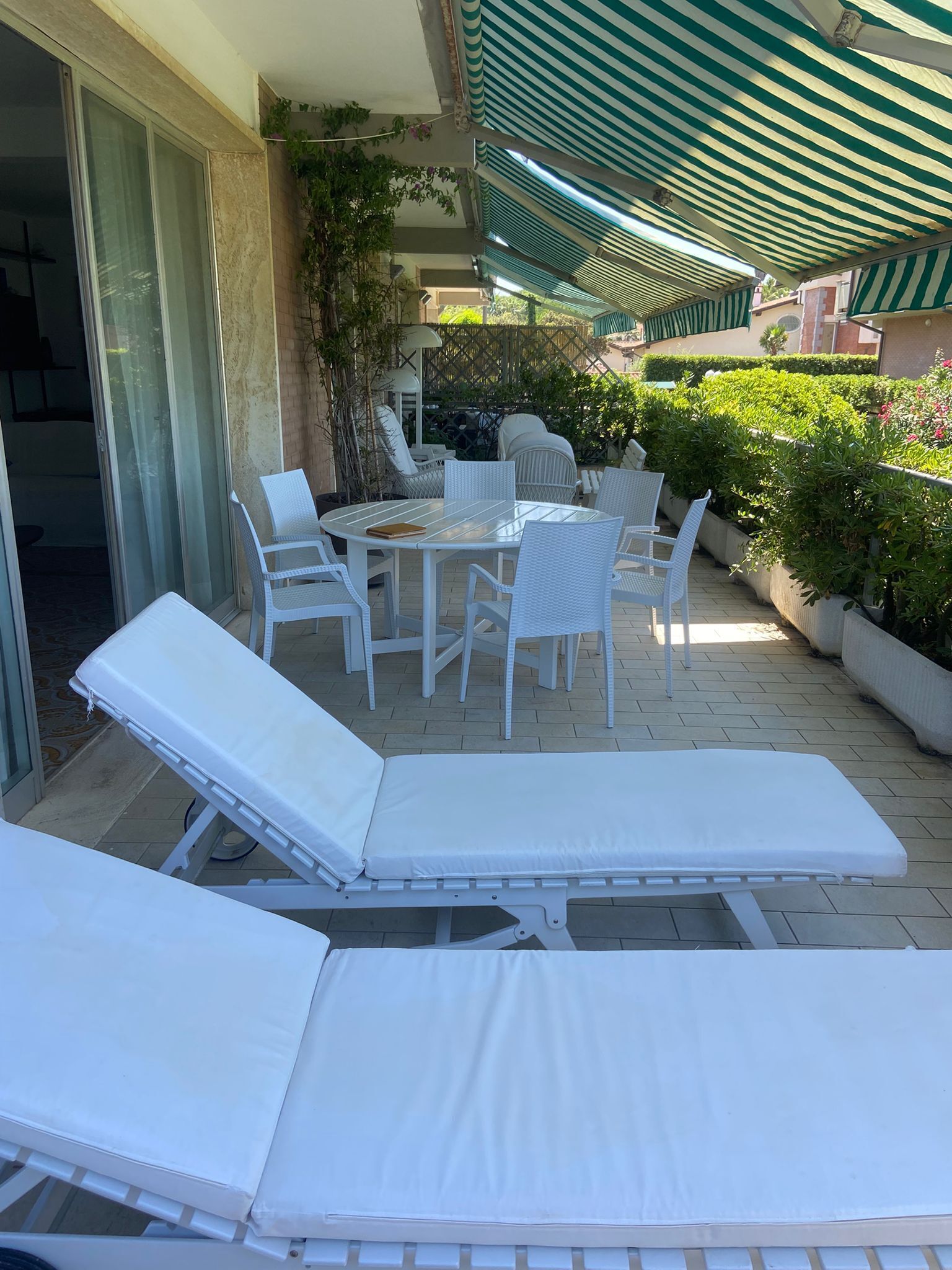 A sunlit outdoor seating area with lounge chairs and a table, surrounded by greenery and a striped awning.