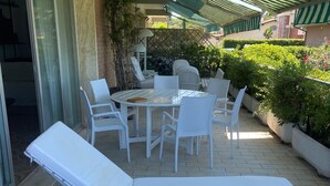 Ein sonnendurchfluteter Außenbereich mit Lounge-Stühlen und einem Tisch, umgeben von Pflanzen und einem Streifenmarkisen.