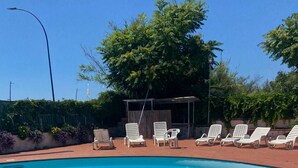 Ein Swimmingpool mit weißen Liegestühlen, ein überdachter Sitzbereich und Bäume im Hintergrund.