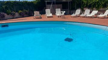 Une piscine entourée de chaises longues blanches, un espace de repos couvert et des arbres en arrière-plan.