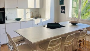 Eine moderne Küche mit weißen Schränken, einer großen Kochinsel mit Barhocker, einer Pendelleuchte und Blick auf Palmen durch das Fenster.