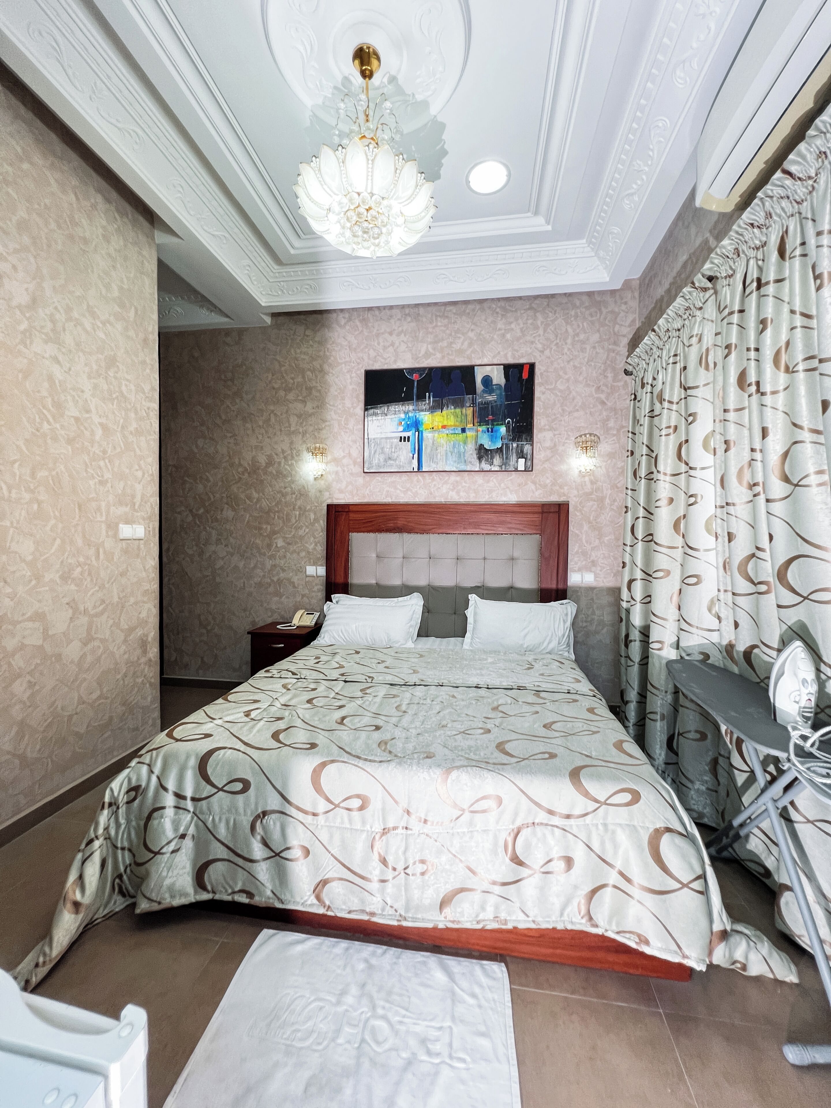 A hotel room with a large bed, a headboard, a nightstand, a telephone, a wall-mounted artwork, and a chandelier.