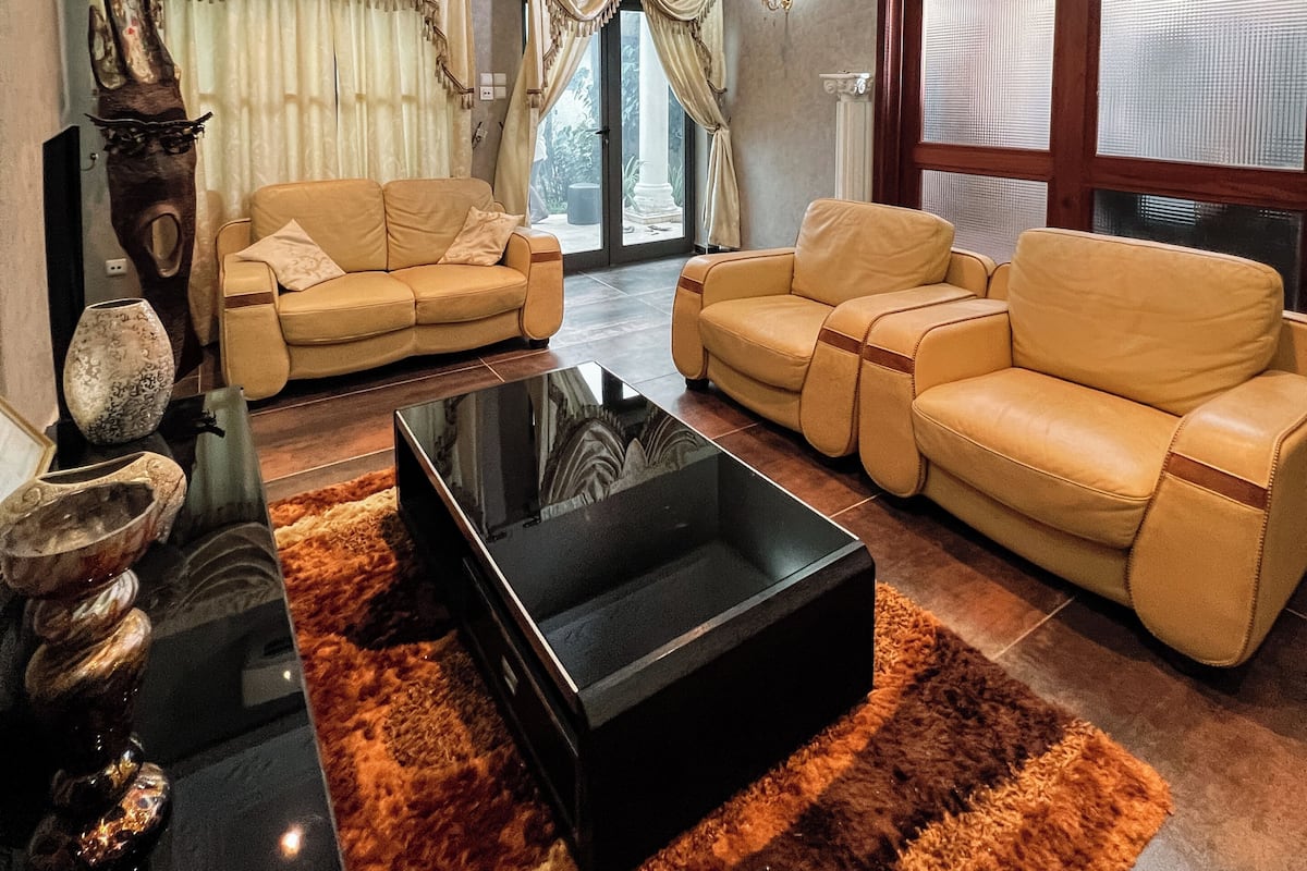 A spacious living room with a large black coffee table, two beige sofas, a chandelier, and a large window with curtains.