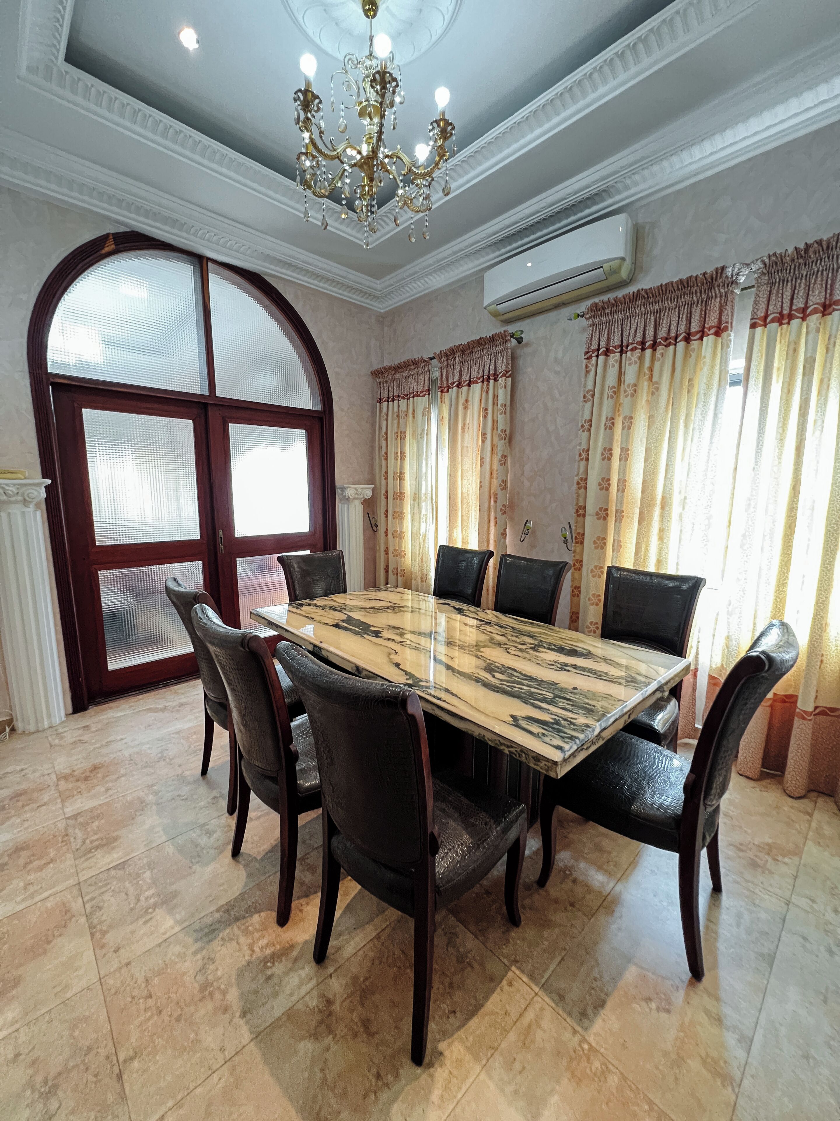 A dining room with a marble table, dark chairs, a chandelier, and large windows with curtains.