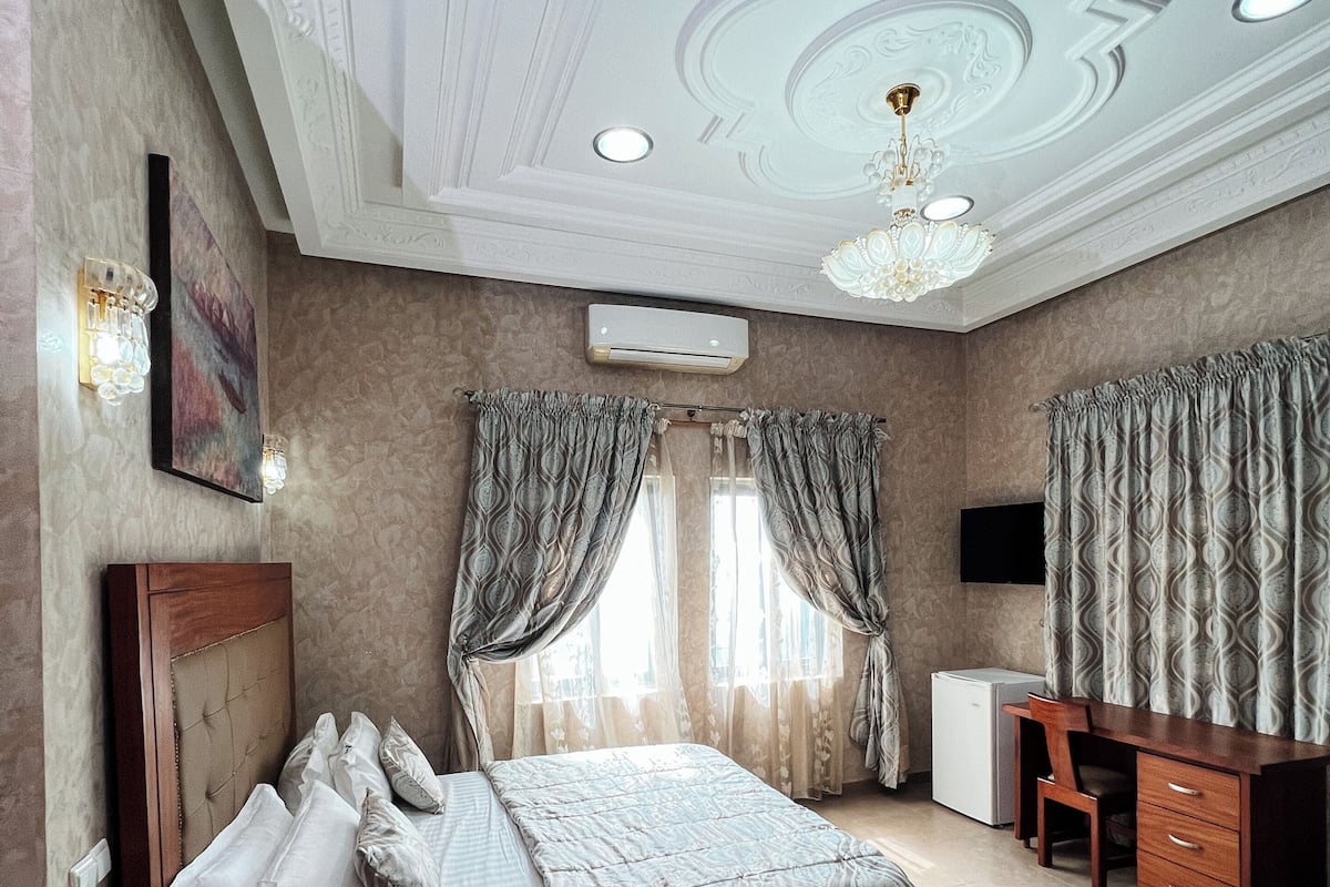 A bedroom with a bed, a desk, a chair, a TV, and an air conditioning unit.