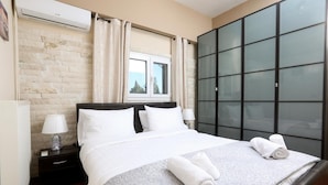A hotel room with a bed, a nightstand, a lamp, a window with curtains, a wardrobe, and a brick wall. - Apartment 'Superior' with Mountain View, Wi-Fi and Air Conditioning (Paleokastritsa)
