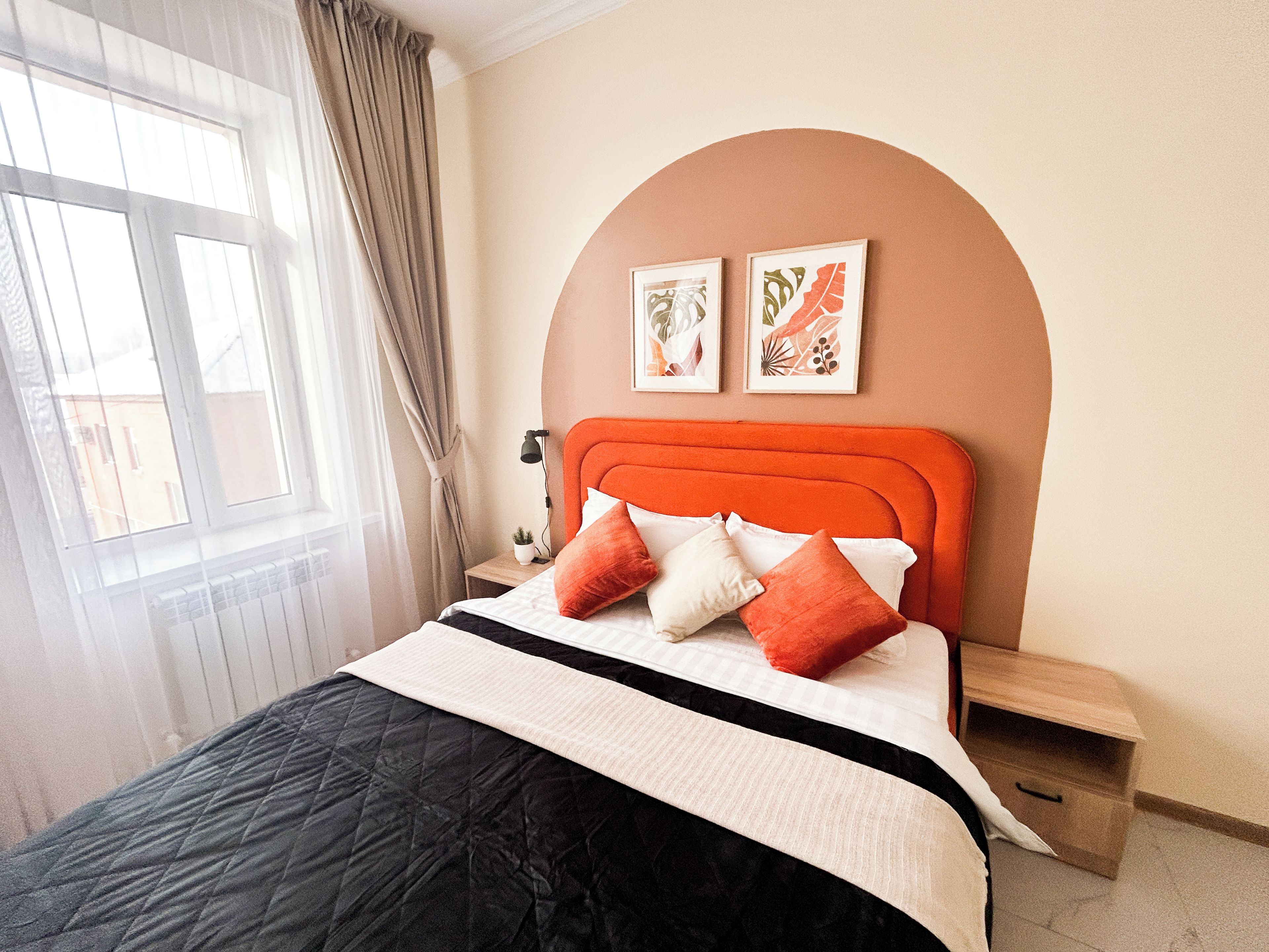 Quarto com cama, mesa de cabeceira, luminária, janela com cortinas e duas imagens penduradas na parede.