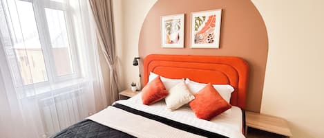 Un dormitorio con cama, mesita de noche, lámpara, ventana con cortinas y dos cuadros en la pared.