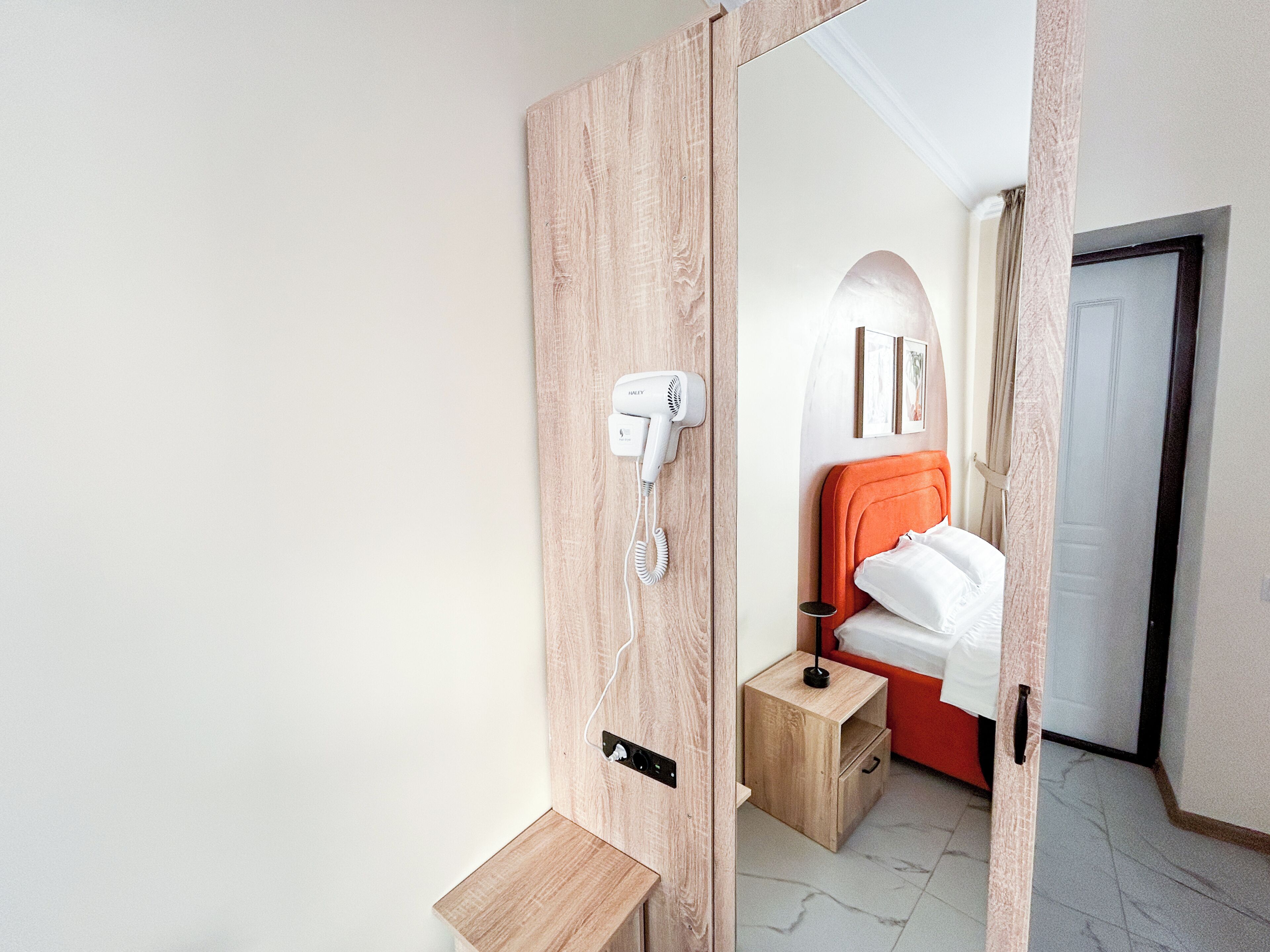Hotellihuone, jossa on puinen ovi, hiustenkuivaaja, pieni puinen yöpöytä, punainen pääty ja valkoinen sänky.