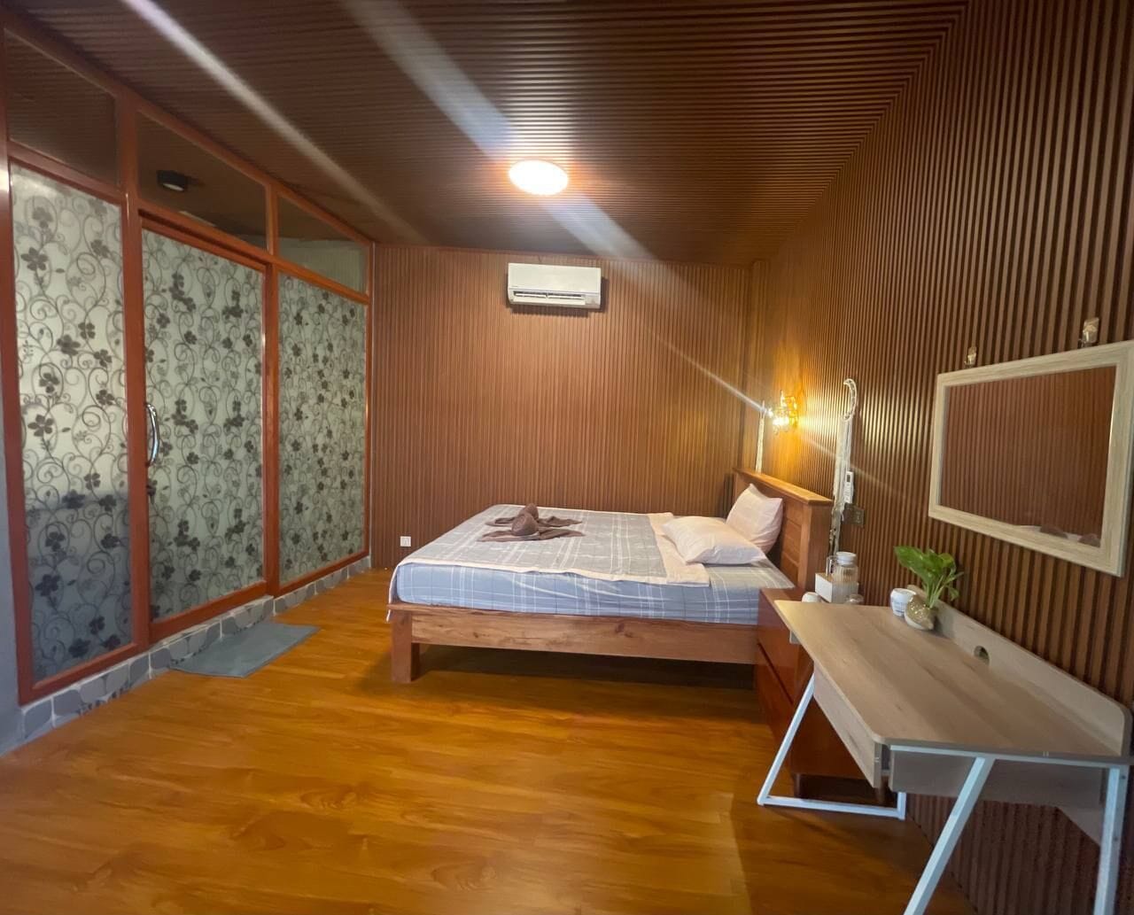 A bedroom with a wooden ceiling, a bed with a blue and white bedspread, a bedside table with a lamp, and a window with patterned curtains.