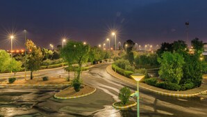 Una carretera urbana nocturna con farolas, una glorieta y zonas verdes.