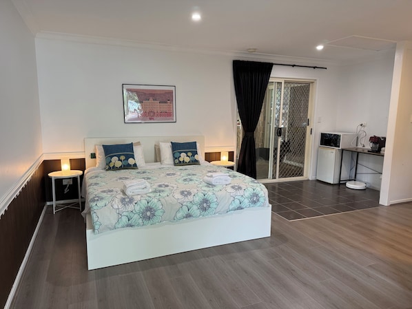 A neatly arranged bedroom with a bed, bedside tables, a small desk, and a sliding glass door leading to a balcony. - Master King Room for Short Stay (Collingwood Park)