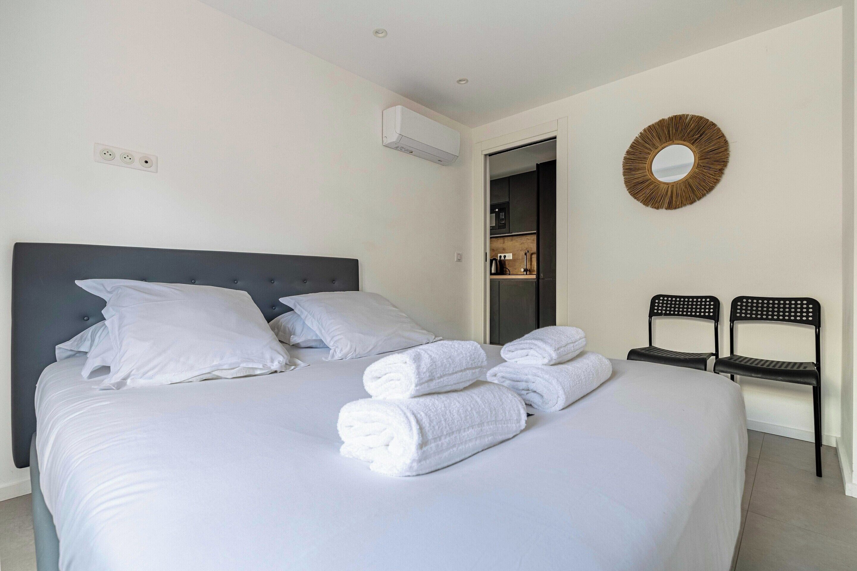 A neatly made bed with white linens and towels, a dark headboard, and a wall-mounted mirror.