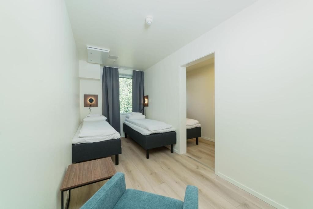 A hotel room with two beds, a small table, a chair, and a window with curtains.