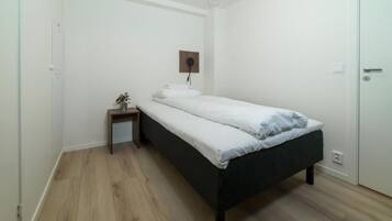 A small, single-bed room with a wooden floor, a bedside table with a plant, and a wall-mounted light fixture.