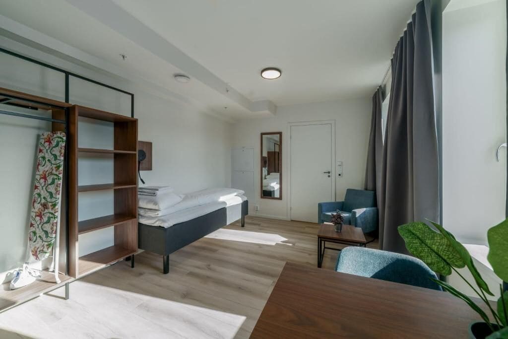 A modern hotel room with a bed, a desk, a chair, a wardrobe, and a mirror.