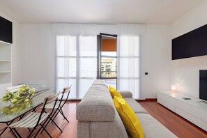 Ein modernes Wohnzimmer mit einer Couch, einem Esstisch mit Stühlen, einem Fernseher und einem großen Fenster.