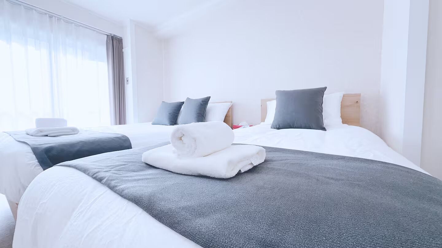 A hotel room with two beds, each with a grey and white bedspread, a wooden headboard, and a window with sheer curtains.