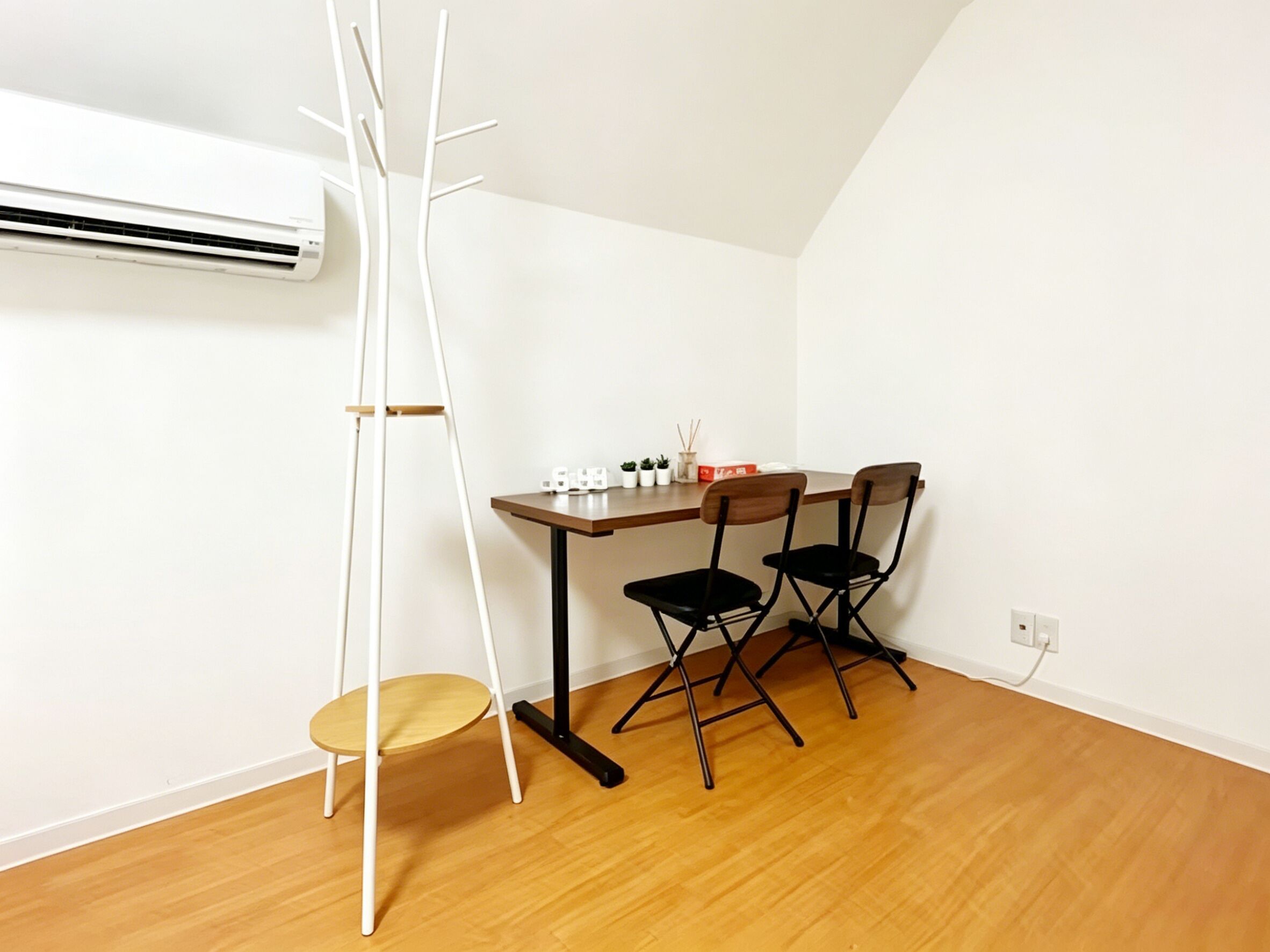 A room with a wooden floor, a white air conditioner, a white ladder, a small table with chairs, and a power outlet.