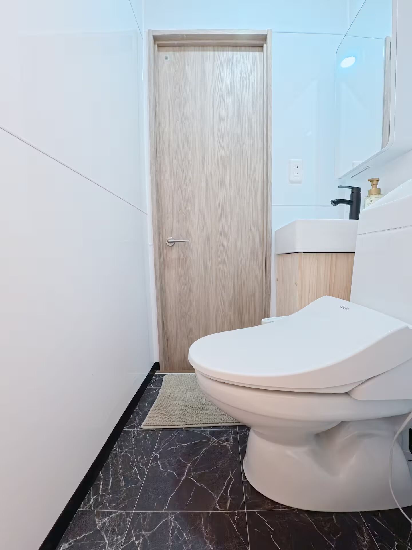 A bathroom with a white toilet, a wooden door, a white sink with a black faucet, and a marble floor.