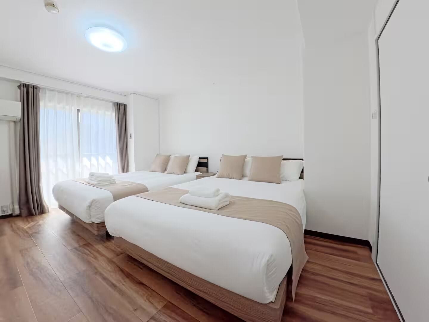 A hotel room with two beds, a large window with curtains, a wooden floor, and a ceiling light.
