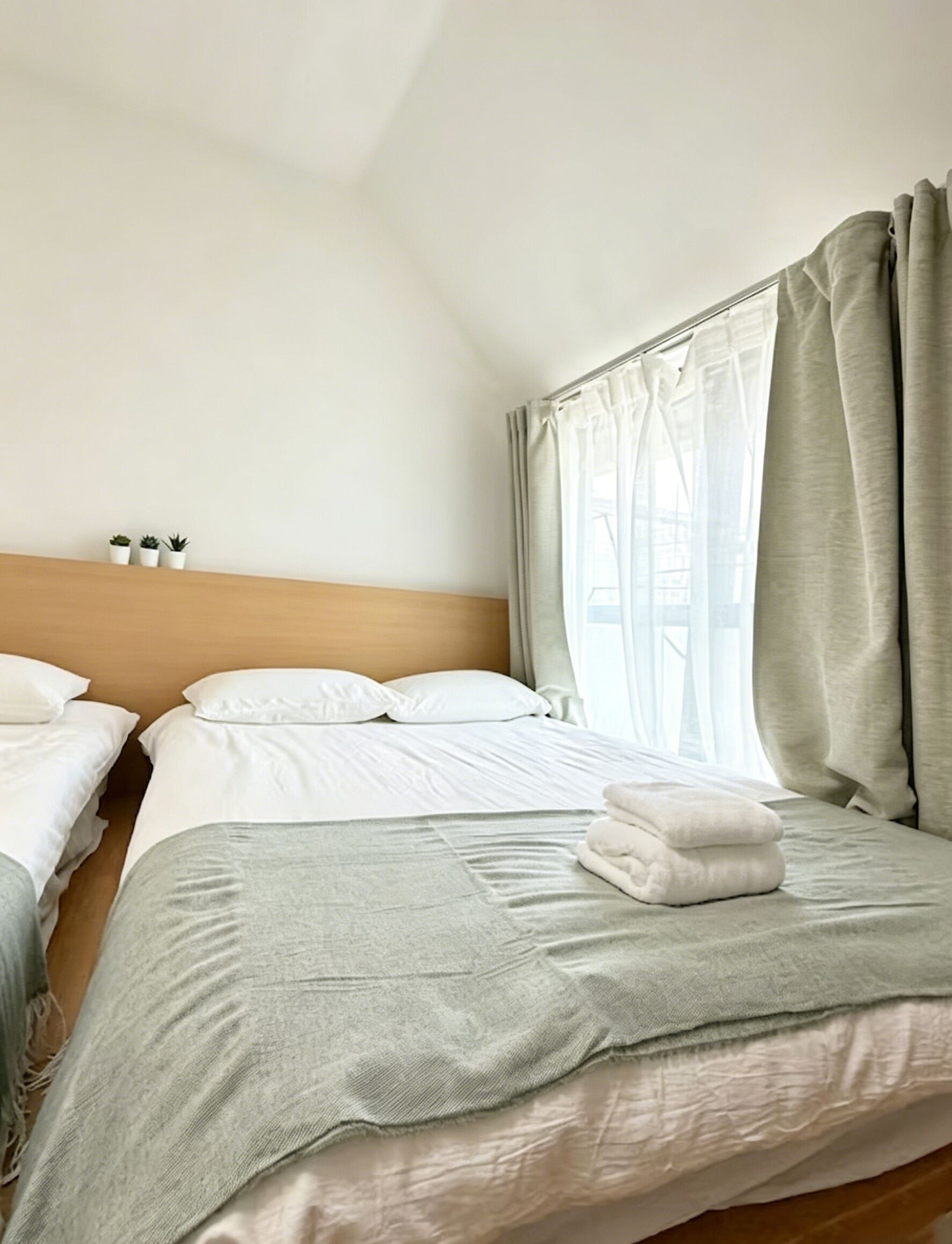 A hotel room with two beds, a wooden headboard, a window with curtains, and a small shelf with bottles.