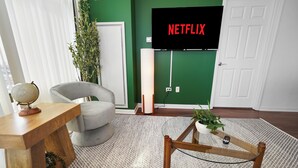 Ein Wohnzimmer mit an einer grünen Wand montiertem Fernseher, einem Globus auf einem Holztisch, einem grauen Sessel und einem kleinen Holztisch.