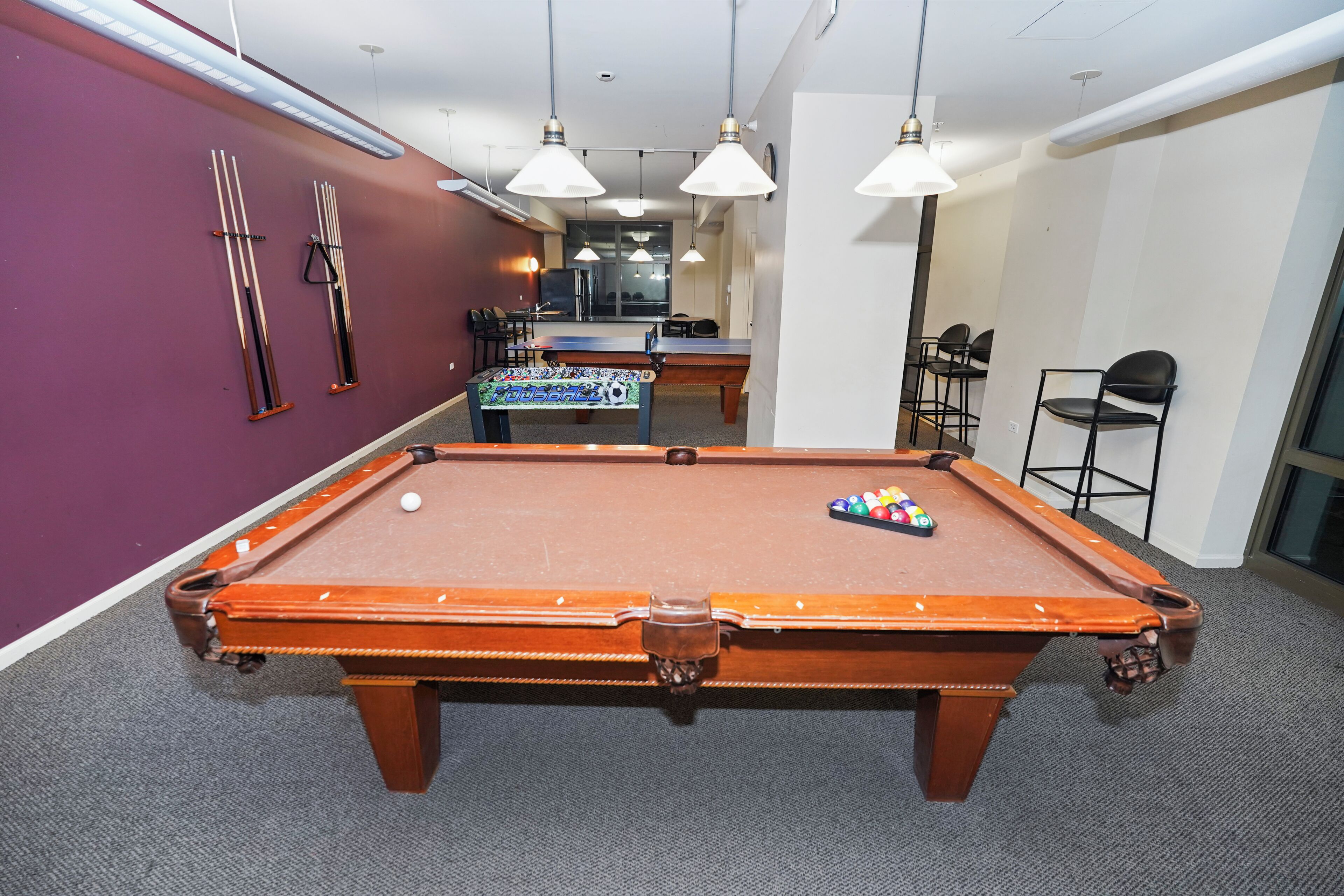A pool table in a room with purple walls, a foosball table, and a bar area.