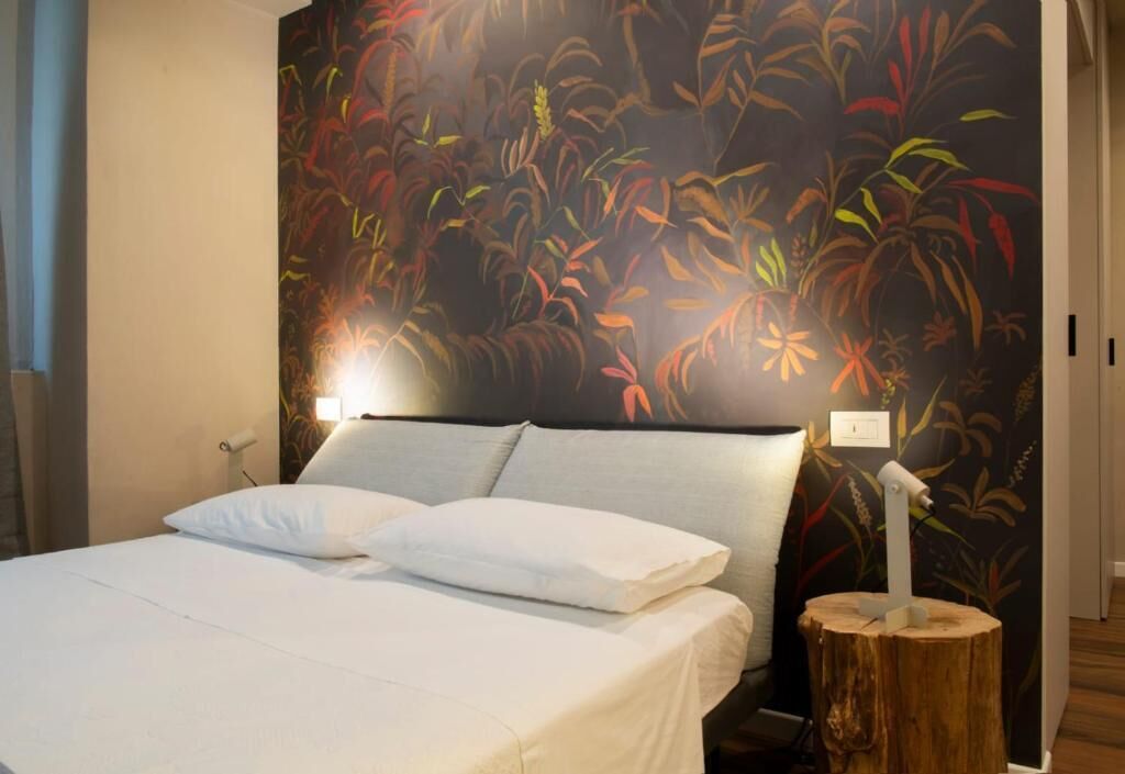 A bedroom with a bed, white bedding, a nightstand, a wall-mounted lamp, and a wooden tree stump.