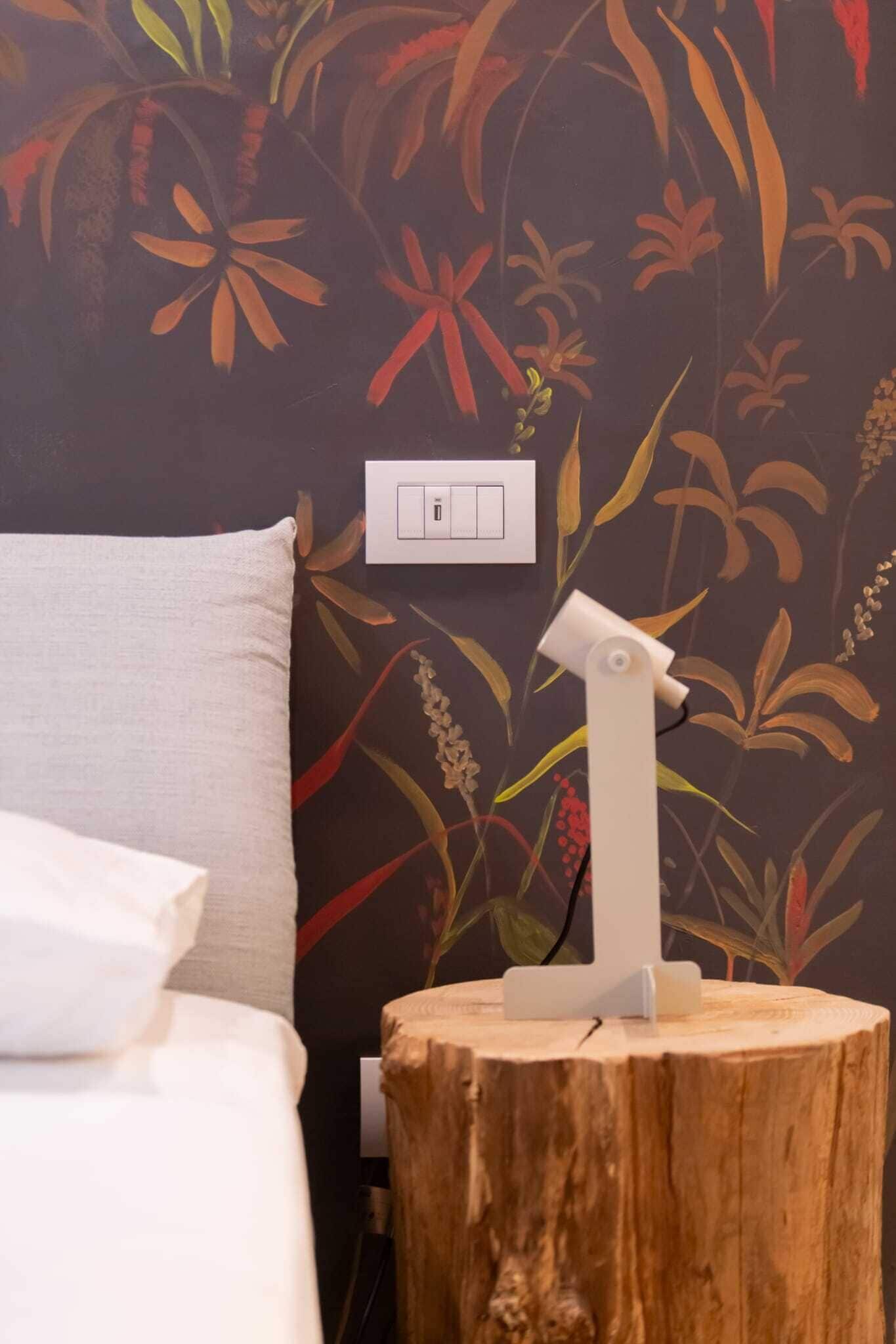 A room with a bed, a white pillow, a wall-mounted light switch, and a wooden stump with a white object on top.