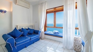 Une pièce avec un canapé bleu, une fenêtre donnant sur la mer, des rideaux blancs et un sol carrelé de couleur blanche.