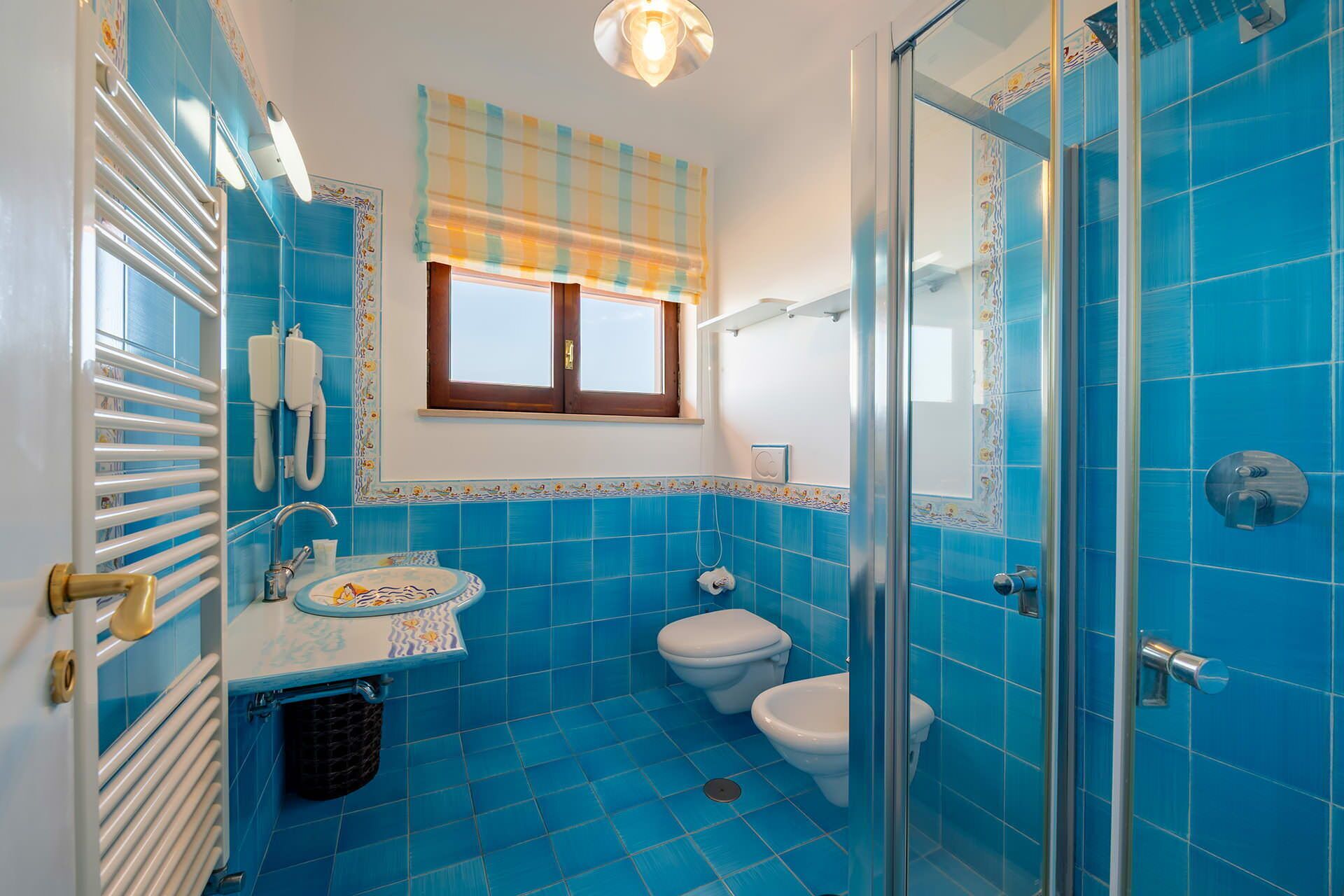 Ein Badezimmer mit blauen Fliesen, einem weißen Waschbecken, einer Toilette, einer Dusche und einem Fenster mit einem Karosmuster-Vorhang.