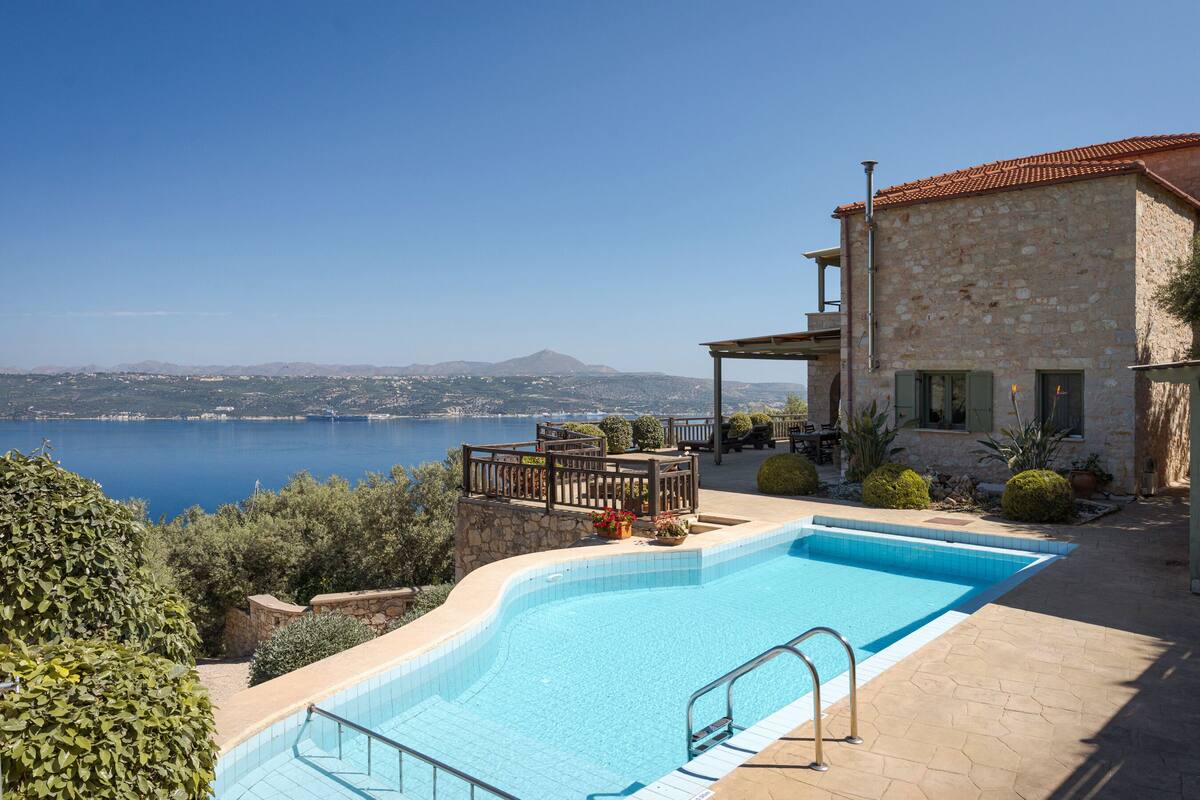A swimming pool with a view of a lake and mountains, a stone building with a tiled roof, and outdoor furniture.