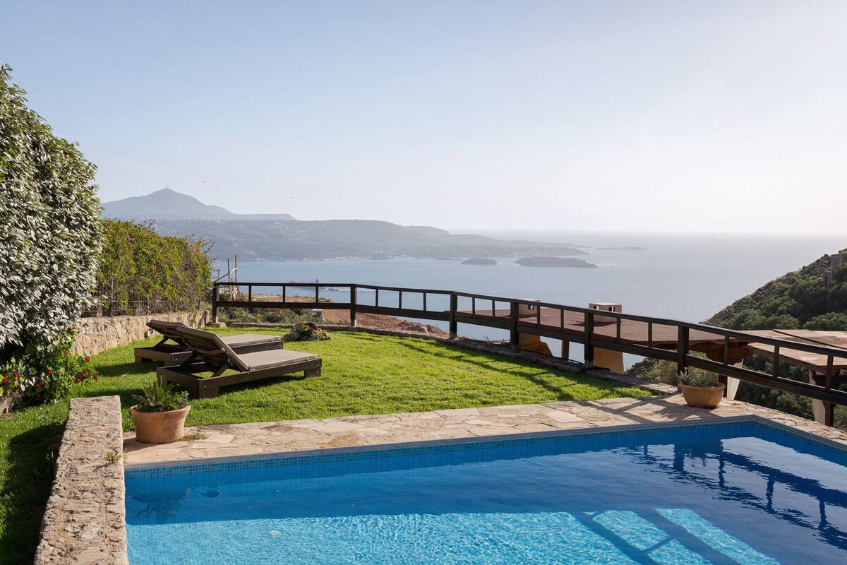 A swimming pool with a wooden deck, a grassy area, and a view of the sea and distant hills.