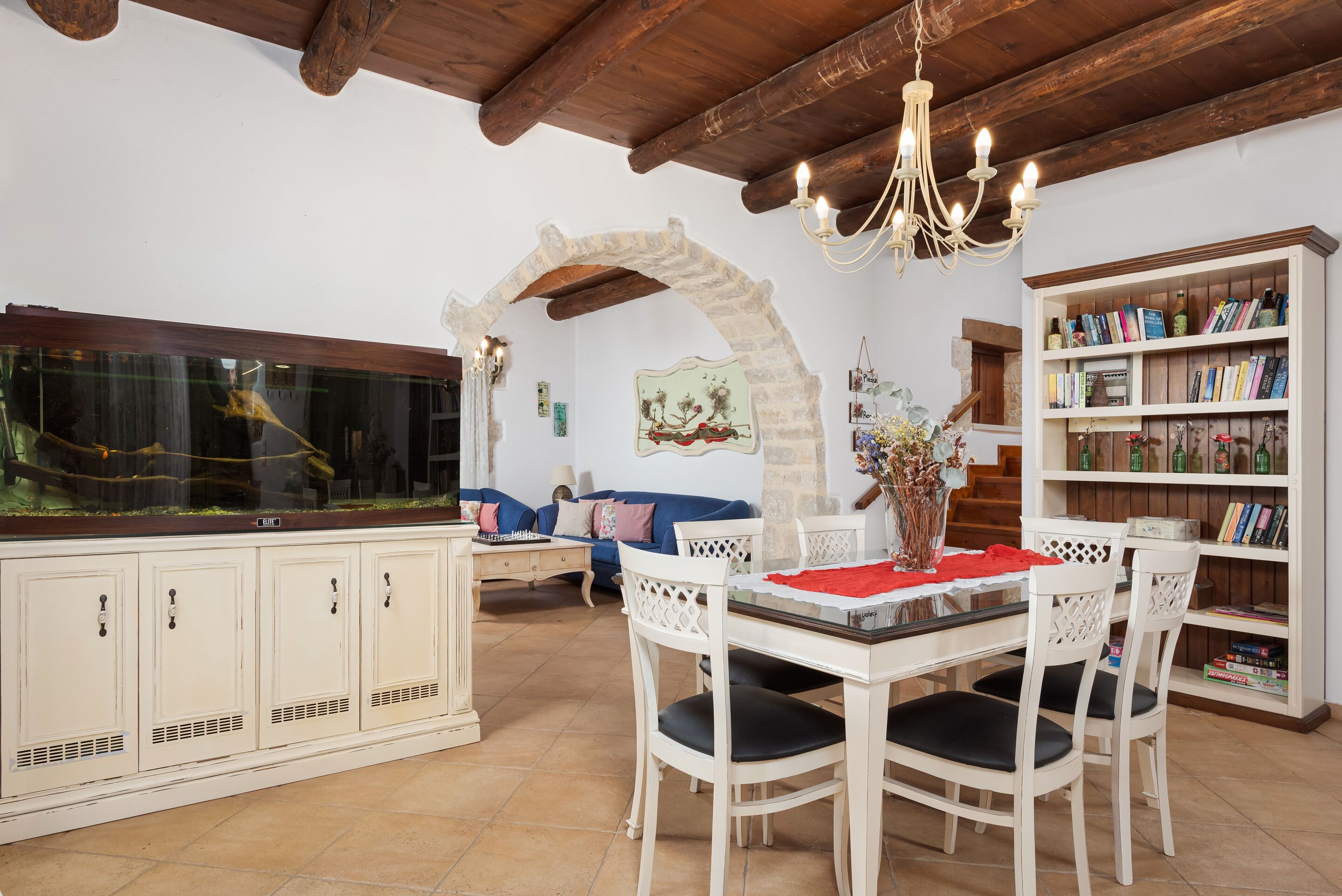 A dining area with a white table, chairs, a chandelier, a bookshelf, and a fish tank.