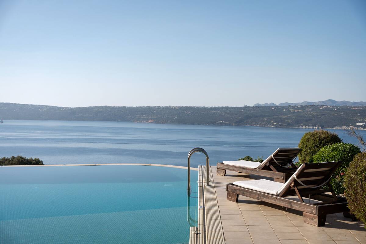 An infinity pool with lounge chairs, a clear view of the sea, and distant hills.