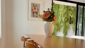 Un comedor con una mesa de madera, sillas, una lámpara colgante tejida, un florero con flores y una obra de arte enmarcada en la pared.