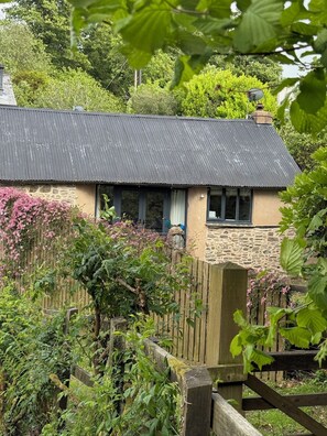 Ein rustikales Steinhaus mit Metalldach, umgeben von üppigem Grün und blühenden Pflanzen.