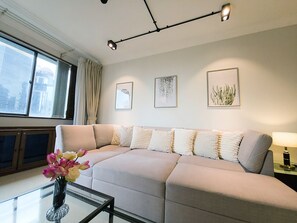 Ein modernes Wohnzimmer mit einer Couch, einem Glastisch und einer Blumenvase. An der Wand hängen gerahmte Kunstwerke und ein Fenster mit Vorhängen.