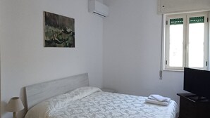 A bedroom with a bed, a chair, a desk, a lamp, a painting, an air conditioner, and a window.