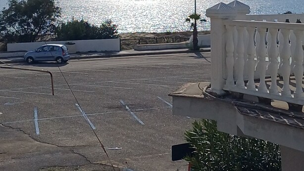 A parking area with a view of the sea, a car, and a building with a balcony.