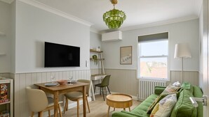 A modern living room with a green sofa, a dining table with chairs, a TV mounted on the wall, a window with a view, and a lamp.